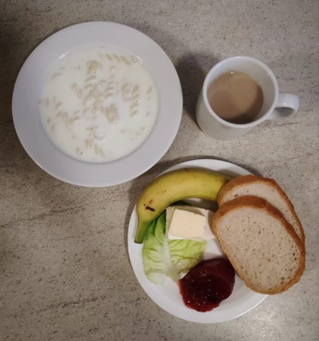 Na stole znajdują się talerz z zupą mleczną, kubek z kawą i talerzyk z sałatą, twarożkiem, dżemem, pieczywo z masłem i banan.