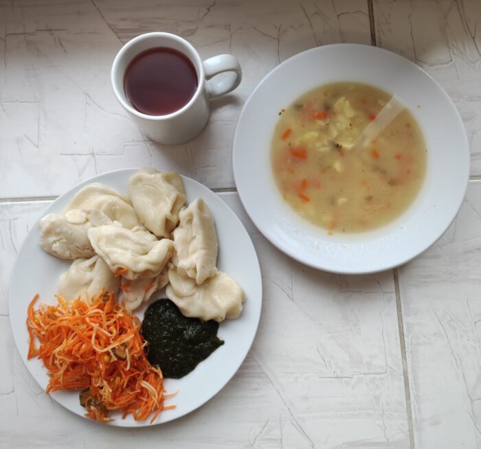 Na stole umieszczone są talerze z zupą, z pierogami, surówką i szpinakiem oraz kubek z kompotem.
