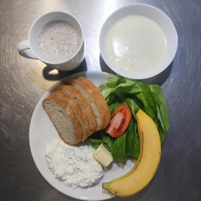 Na stole znajdują się talerz z zupą mleczną, kubek z kawą i talerzyk z twarożkiem, sałatą, pomidorem, bananem i pieczywo z masłem