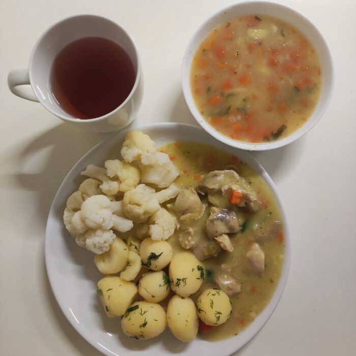 Na stole znajduje się talerz z zupą koperkową, kubek z kompotem i talerz z kalafiorem ziemniakami i gulaszem drobiowym