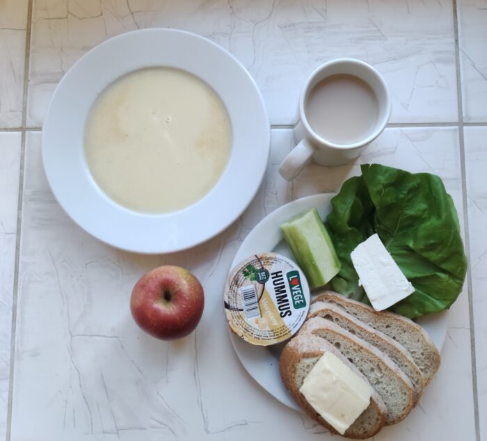 Na stole są talerze z zupą mleczną, chlebem, masłem, twarogiem, hummusem i warzywami oraz kubek z kawą i jabłko