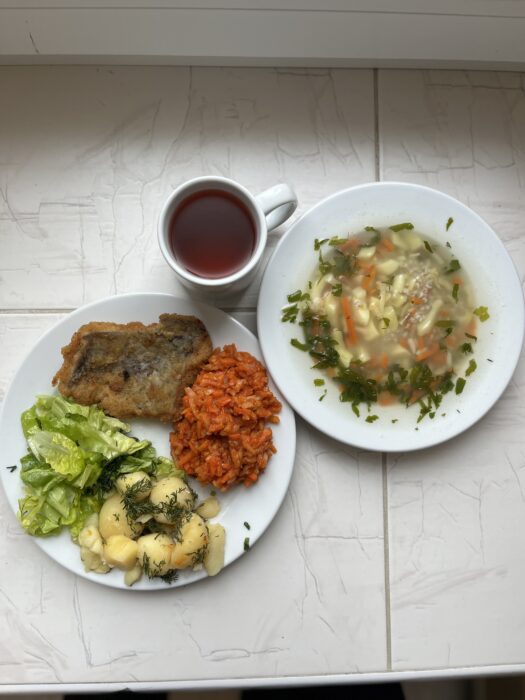 Na stole są talerze z zupą, rybą, ziemniakami, marchewką, sałatą oraz kubek z kompotem.